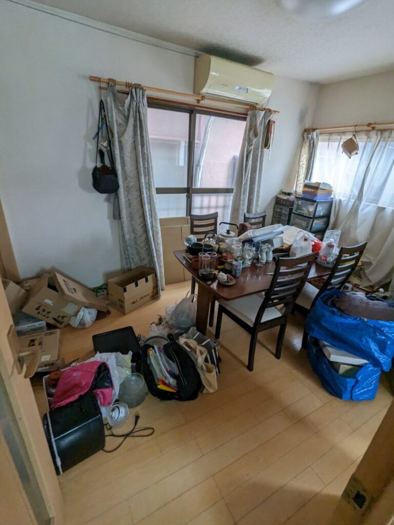共有持分トラブルで買取した物件の室内。テーブルや床に荷物や段ボールが散らかったままの居室で、窓際にはカーテンと古いエアコンが設置されている。
