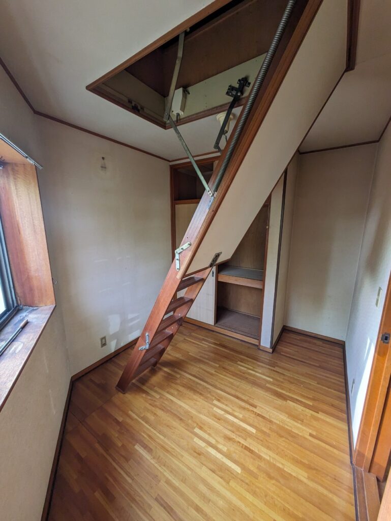 折りたたみ式の天井収納ハシゴが下りた室内の様子。フローリングの床と壁の劣化が見られ、奥には収納棚が備わっている。