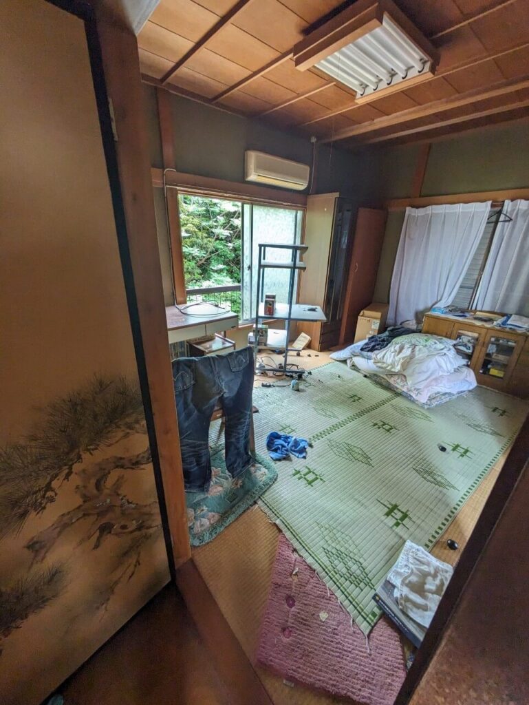 残された布団や衣類、家具があり、畳の劣化が目立つ空き家の和室内部の様子を写した写真