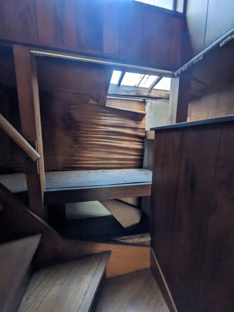 老朽化が進んだ木造住宅の階段周り。壁材が大きく波打ち、上部の天井も破損して光が差し込むなど、内部の損傷が目立つ状態。