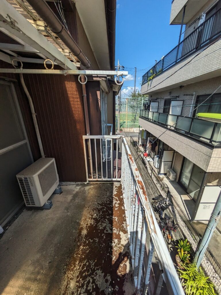 老朽化が進んだバルコニーの様子。床面や手すりに錆びや劣化が見られ、隣接する建物との距離が近い住宅環境が分かる。