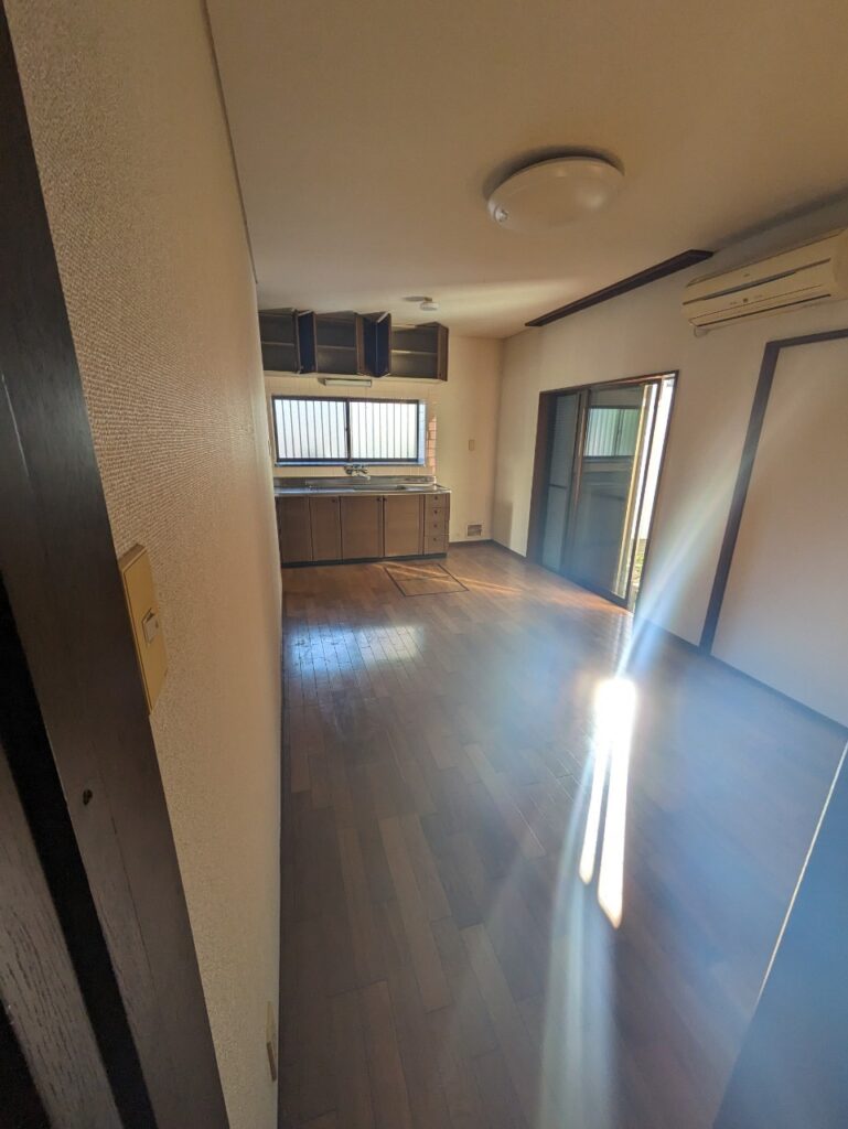 窓から日差しが差し込む室内。経年劣化したフローリングと古いキッチンがあり、相続後放置されていた連棟住宅の内部が分かる。