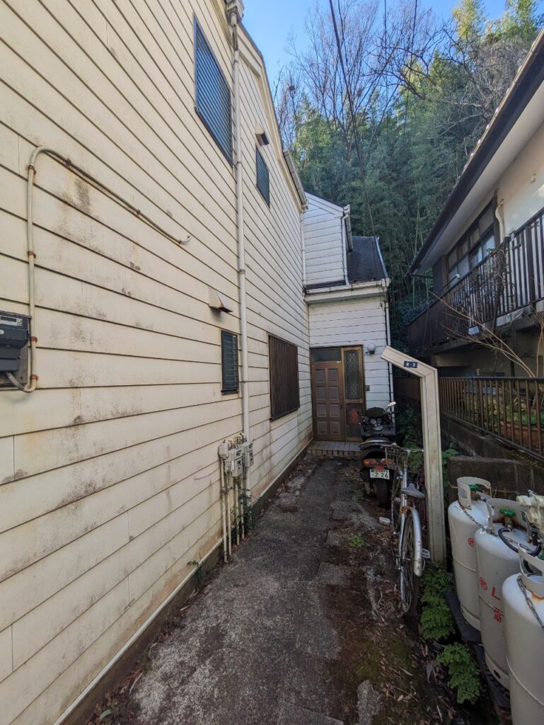 連棟住宅の裏側通路の様子。白い外壁には汚れや劣化が見られ、細い通路には自転車やバイクが置かれている。