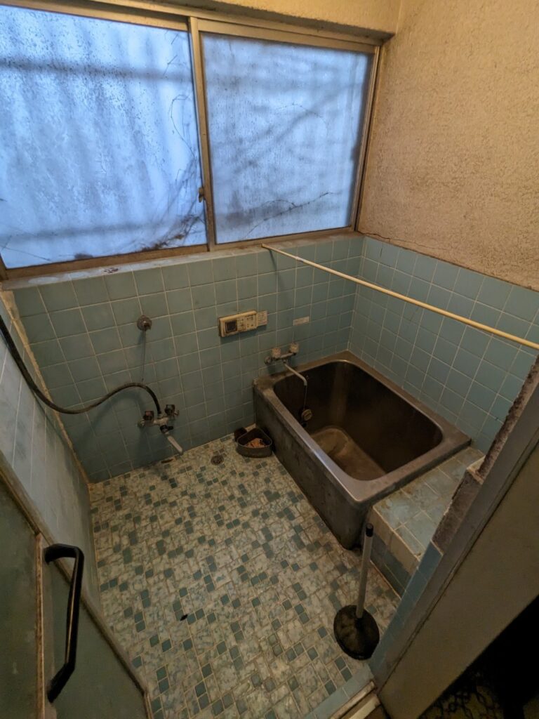 劣化が進んだ浴室の様子。曇った窓と青いタイル張りの壁・床、ステンレス浴槽があり、長期間使用されていなかった状態がうかがえる。