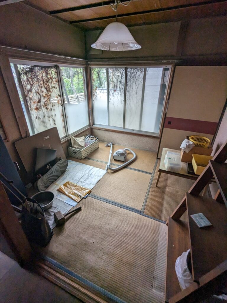 長期間放置され劣化が進んだ和室の様子。汚れた障子付きの窓や傷んだ畳があり、室内には掃除機や工具などの荷物が残された状態。