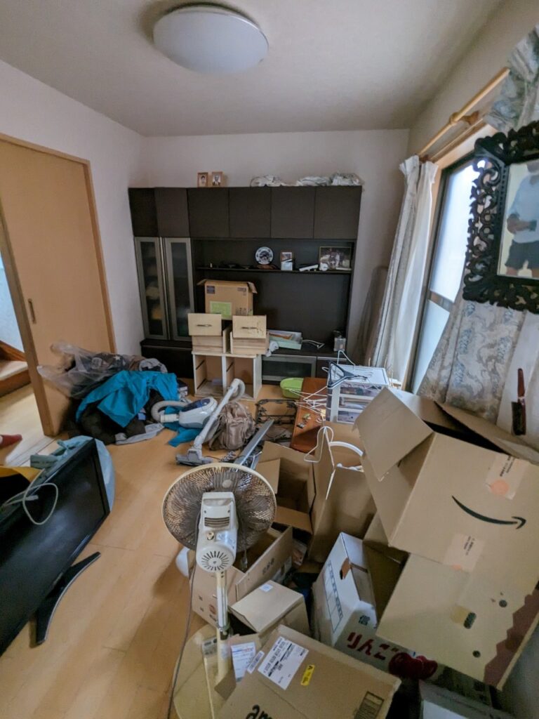 荷物や段ボールが散乱した状態の居室。棚や家電、衣類などがそのまま残置されており、共有持分トラブル物件として買取した際の室内状況が分かる。