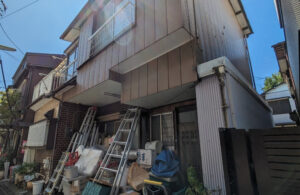世田谷区鎌田で買取した再建築不可物件の外観。幅員約1mの細い通路に面し、家の前には荷物や脚立が積まれたままの空き家の様子。
