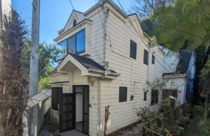 横浜市港南区日野で買取。相続後に放置されていた連棟住宅の外観。細い通路沿いに建ち、経年劣化が見られる白い外壁と黒い玄関扉が特徴の空き家。