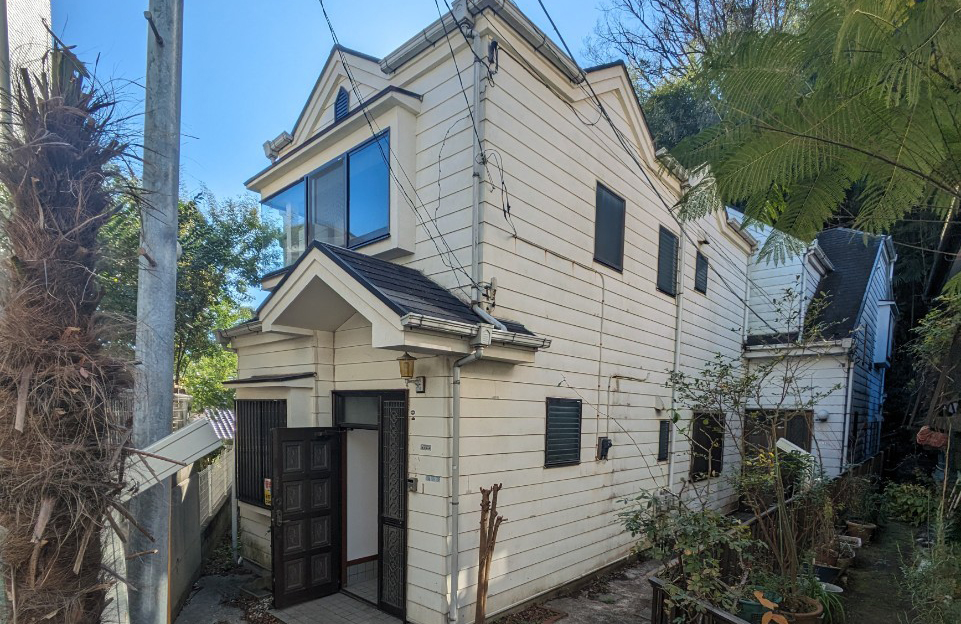 横浜市港南区日野で買取。相続後に放置されていた連棟住宅の外観。細い通路沿いに建ち、経年劣化が見られる白い外壁と黒い玄関扉が特徴の空き家。