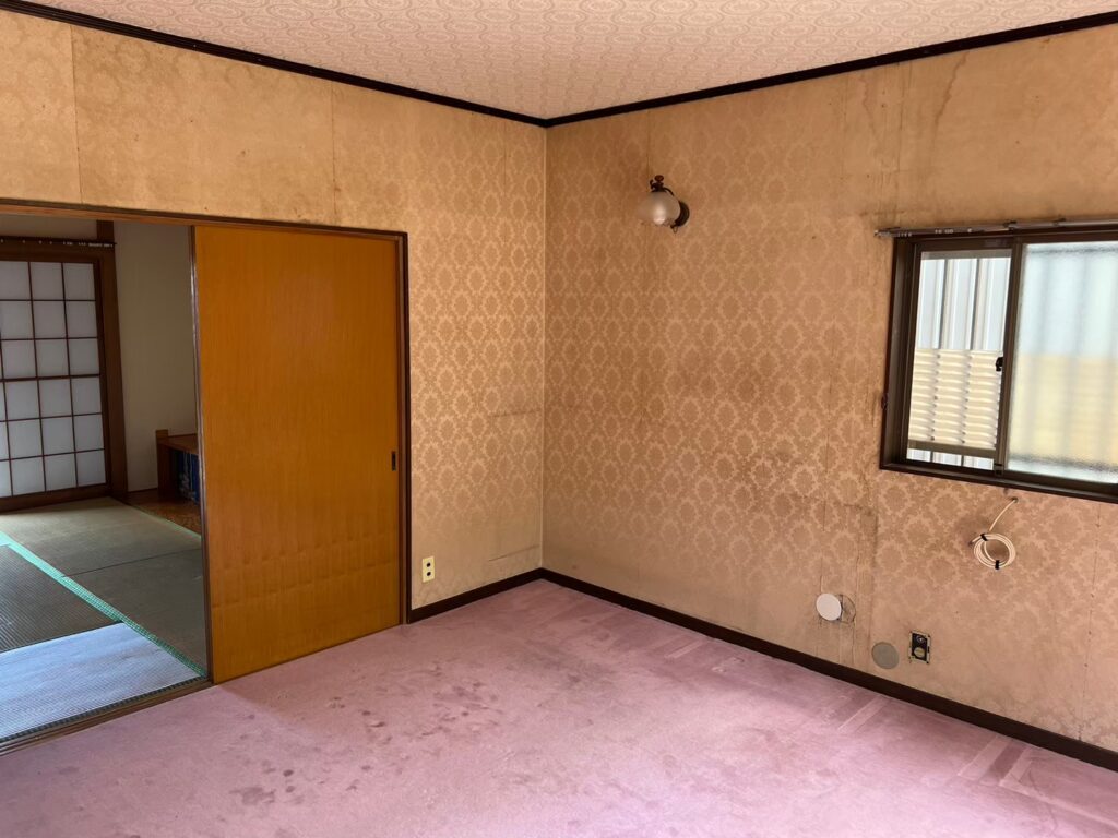 室内の壁紙に劣化や汚れが見られる居室の様子。ピンク色のカーペットが敷かれ、左奥には襖と畳の和室が続いている。