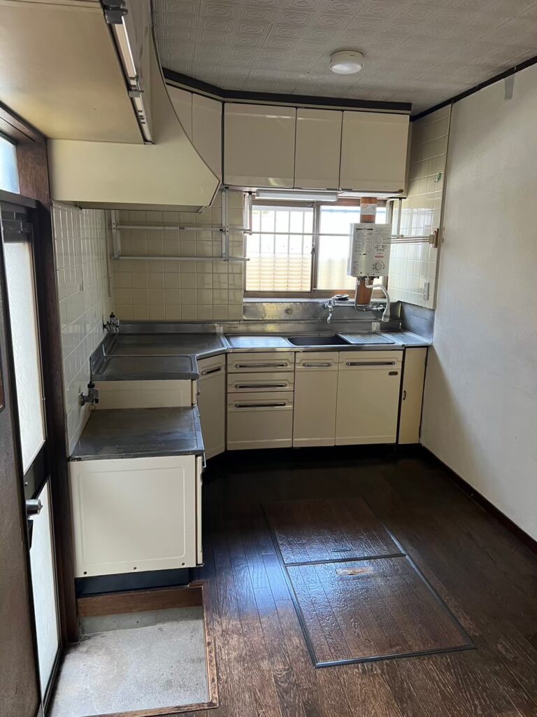 経年劣化が見られるキッチンの室内写真。吊り戸棚やステンレスの調理台、給湯器が備え付けられており、床下収納のあるフローリング。